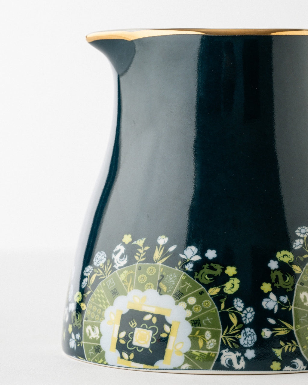 Navy ceramic milk pitcher with curved spout and gold-trimmed rim, displaying green and white botanical pattern on lower portion against white background