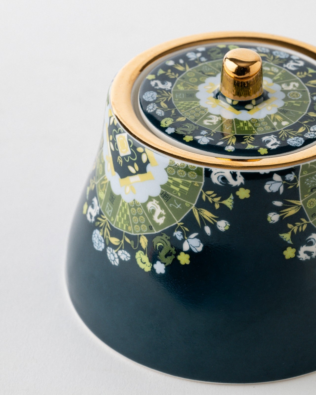 Close-up of navy ceramic lidded sugar bowl with gold-trimmed lid, brass finial, and green botanical pattern detail on white background