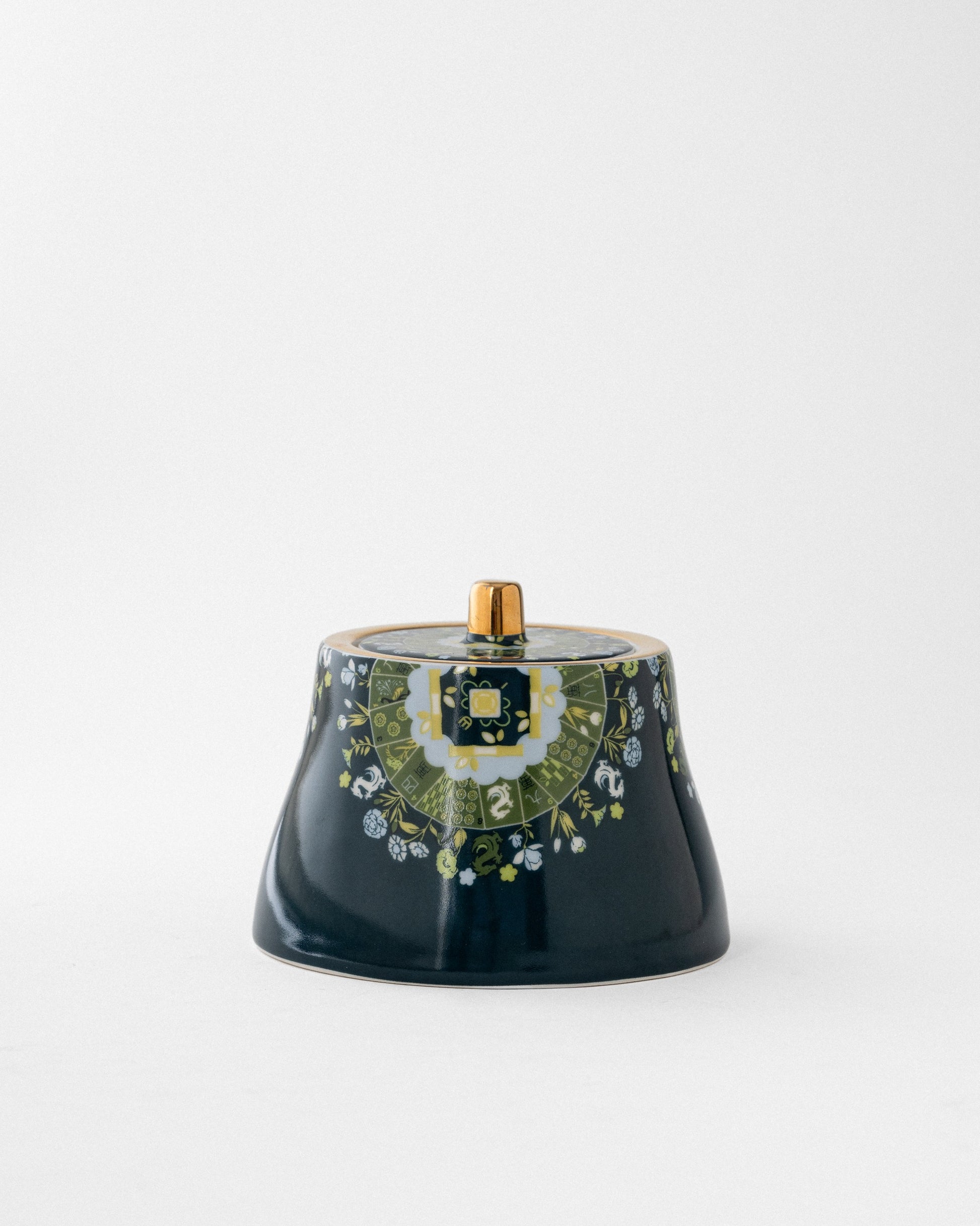 Navy ceramic covered sugar vessel with brass finial, featuring green and white botanical border band beneath gold-trimmed lid on neutral background